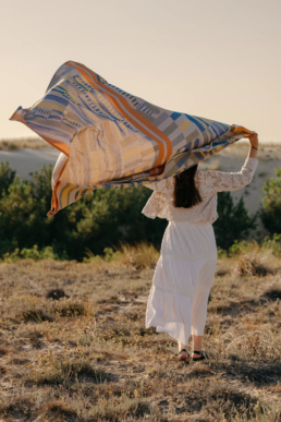 emme de dos vêtue d'une robe blanche longue tenant au vent une grande nappe colorée Tissage Moutet dans un paysage de dunes.