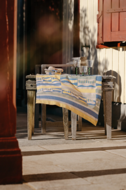 Mise en scène d'une table avec du linge de maison jacquard Tissage Moutet, trois verres texturés et un coquillage sur une terrasse en bois.
