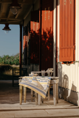 Petite table en bois rustique dressée avec une nappe Tissage Moutet sur la terrasse ombragée d'une cabane aux volets rouges.